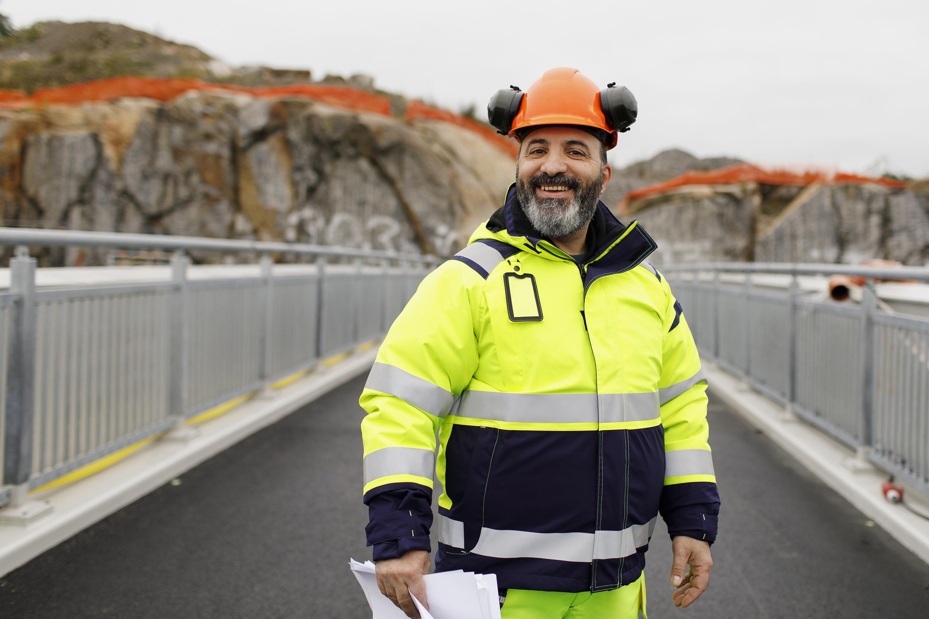 En person i reflexjacka, skyddshjälm och hörselskydd står på en bro och ler mot kameran. Hen håller i papper och har berg i bakgrunden
